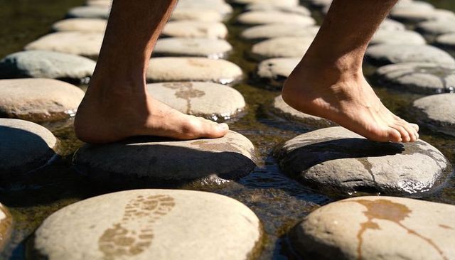 Barefoot adult crossing round stepping stones in sunlit shallow stream