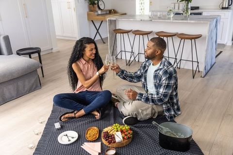 Couple clinking wine glasses at romantic indoor picnic with cheese board and ice bucket