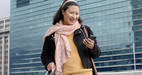 Mature Woman Walking in Plaza Wearing Pink Scarf Checking Phone Carrying Shopping Bags