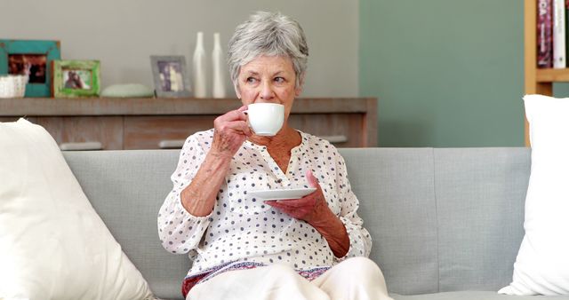 Senior woman sitting on light gray couch, sipping tea from white cup with saucer. Casual and relaxed setting, home interior. Ideal for themes related to elderly lifestyle, relaxation at home, and peaceful living. Suitable for use in blogs, advertisements, and articles focused on senior living, home comfort, and leisurely activities for older adults.