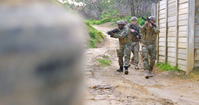 Three soldiers in camouflage uniforms and helmets performing a combat training drill with rifles. They are in a tactical stance, moving along a dirt path, prepared and focused on their mission. Ideal for use in military training manuals, defense force promotional materials, and articles on military readiness and teamwork.