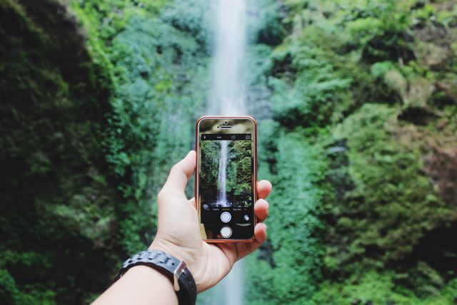 Hand holding smartphone with lush, green waterfall scenery in background, ideal for content about travel, outdoor adventures, nature photography, and technology usage in tourism.