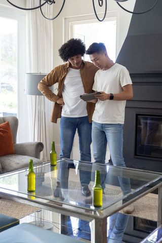 Diverse young men leaning and examining tablet in modern living room with green bottles