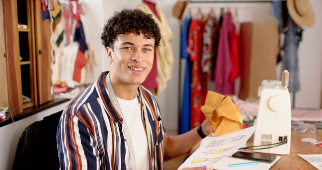 Happy biracial male fashion designer sitting by desk and holding fashion designs in studio. Fashion, design, creativity, clothing and small business, unaltered.