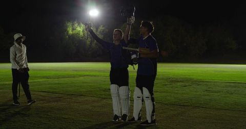 Night cricket celebration under floodlights two batsmen raising helmets after century