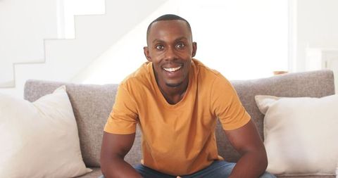 Smiling young man leaning on sofa wearing orange t-shirt in bright modern living room