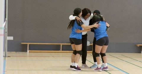 Coach celebrating with diverse female volleyball team huddling near net on indoor gym court