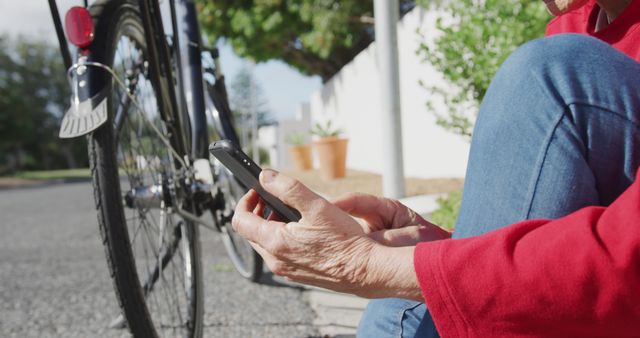 Senior person sitting near a bicycle, using a smartphone on a sunny day. Ideal for depicting active aging, technology use among seniors, urban transportation, and leisure activities in outdoor settings.