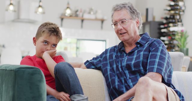 This image depicts an older man and a young boy sitting on a couch in a cozy living room. The grandfather is wearing a blue plaid shirt and appears to be engaging in conversation, while the boy looks bored and is resting his chin on his hand. This photo can be used in articles or advertisements about family relationships, generational differences, home life, and bonding moments. It is perfect for illustrating themes of family interaction and communication within a domestic setting.