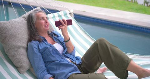Mature woman relaxing on poolside hammock speaking into smartphone
