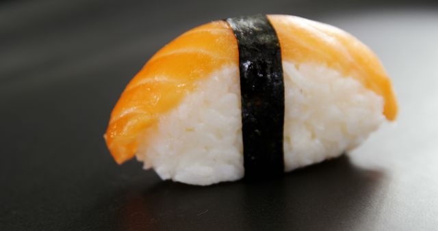 A piece of nigiri sushi, featuring a slice of fresh salmon atop a bed of vinegared rice wrapped with a strip of nori, is presented on a dark surface. Sushi is a traditional Japanese dish enjoyed worldwide for its delicate flavors and artistic presentation.