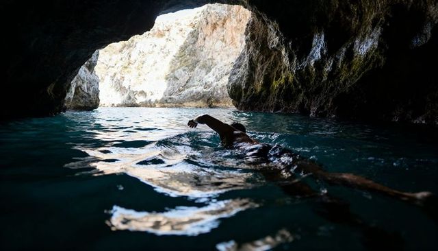 Swimmer paddling toward sunlit sea cave with turquoise water, rocky arch and reflections