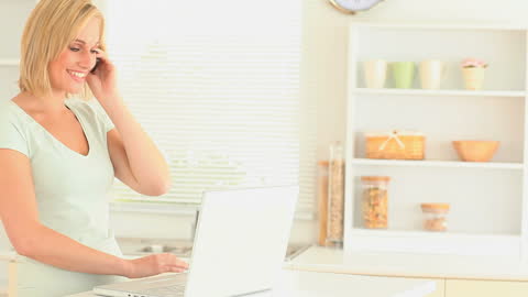 Woman Speaking on Phone While Using Laptop in Kitchen