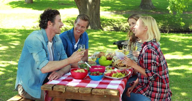 Group of friends enjoying picnic in park on sunny day, smiling and chatting around table filled with food and drinks. Perfect for promoting outdoor activities, social gatherings, and healthy lifestyle.