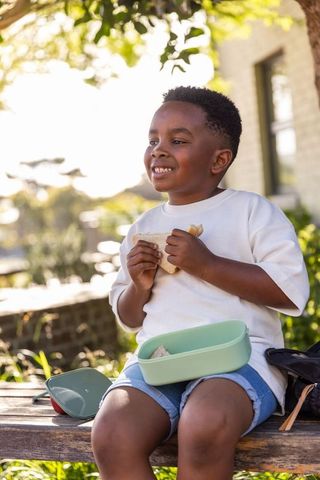 Sunlit african american boy enjoying sandwich on wooden bench with mint green lunchbox