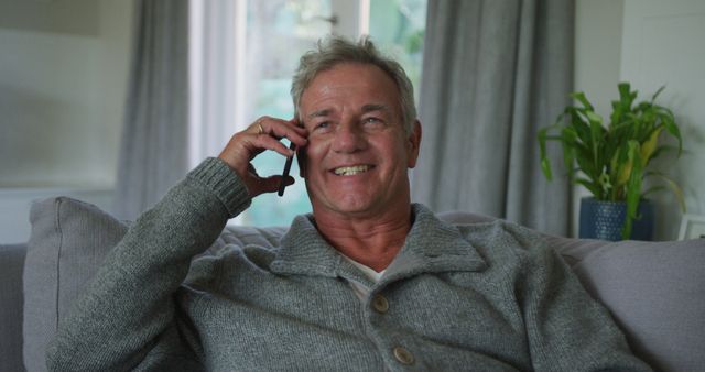 Mature man in comfortable home setting, wearing a gray sweater, enjoying a phone conversation while sitting on a sofa. Can be used for themes related to communication, senior lifestyle, relaxation, domestic settings, and friendly interactions.