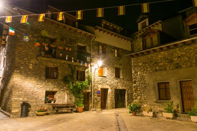 This scene captures a charming medieval village at night with stone buildings warmly illuminated by streetlights. The ambient night Ssceneries perfect for use in travel websites, brochures, marketing materials, or cultural presentations about European history and architecture. The hanging flags add a touch of local tradition, making this image suitable for features on historical destinations or small-town charm.
