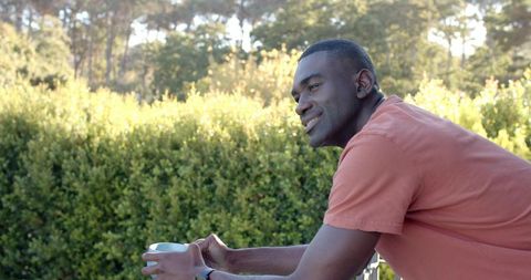 African American man relaxing on balcony holding mug leaning on railing in sunlit garden