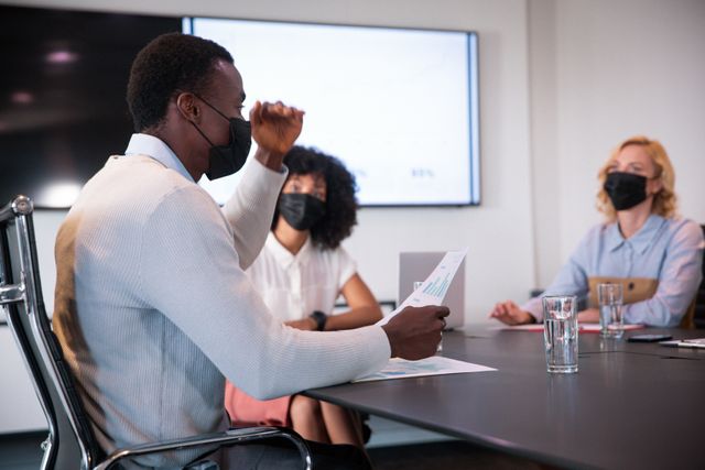 Business professionals wearing face masks are engaged in a discussion in a modern office setting. This image can be used to illustrate themes related to workplace safety, teamwork, and business operations during the COVID-19 pandemic. It is suitable for articles, presentations, and marketing materials focusing on health precautions, corporate environments, and pandemic-related adjustments in the workplace.