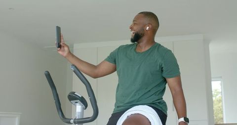 Smiling man cycling on stationary bike at home using smartphone and earbuds