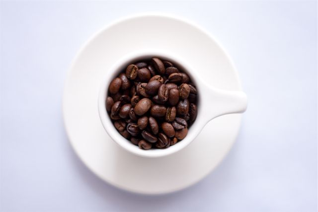 Capturing coffee culture, this image shows a white cup filled with richly roasted coffee beans on a matching saucer. Perfect for use in coffee shop promotions, gourmet coffee blogs, beverage articles, or as a feature image for cafe menus. It emphasizes simplicity and the aromatic appeal of fresh coffee beans.