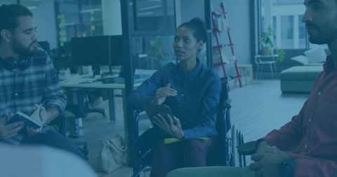 Woman in wheelchair leading meeting while holding tablet in modern open-plan office