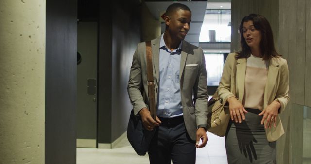 Two business professionals, a man and a woman, walking and engaging in conversation in an office corridor. Both are wearing business attire and carrying bags, suggesting a setting suitable for corporate use. This can be used in articles or websites that discuss business communication, workplace diversity, casual meetings, and office culture.