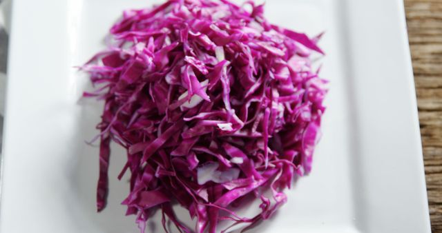 Fresh shredded red cabbage neatly arranged on a square white plate. Ideal for use in articles about healthy eating, vegetarian recipes, food blogging, cooking tutorials, and nutritional guides. Highlights the vibrant color and raw texture, making it suitable for visually appealing salads and vegetarian dishes.