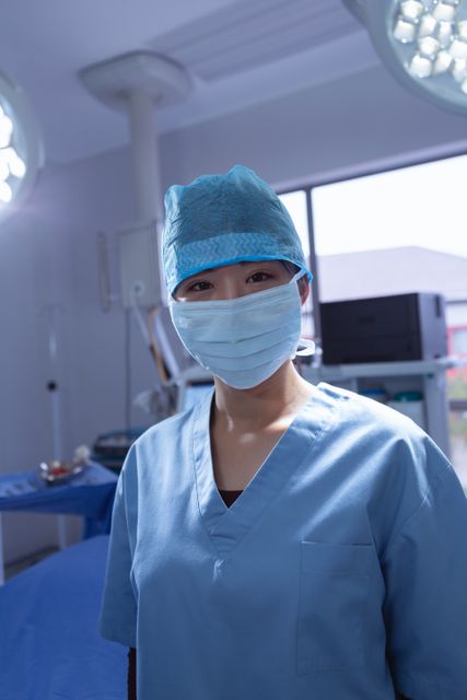 Image depicts a female surgeon wearing a surgical mask and scrubs, standing in an operation room. She is looking directly at the camera, suggesting readiness and confidence. This image can be used in healthcare-related content, medical websites, hospital brochures, and educational materials to illustrate surgical procedures, healthcare professionals, and hospital environments.