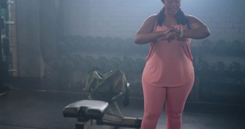 African american woman checking wrist tracker in gym wearing pink activewear near bench