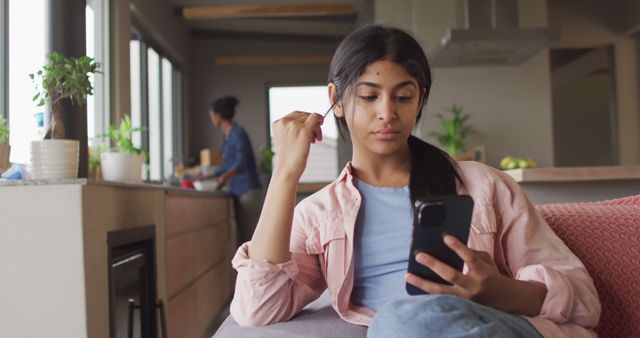 Image of thoughtful biracial woman sitting on sofa and using smartphone. Relax, leisure and spending free time at home with technology concept.