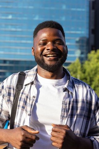 Confident african man smiling in urban business district wearing checked shirt and backpack