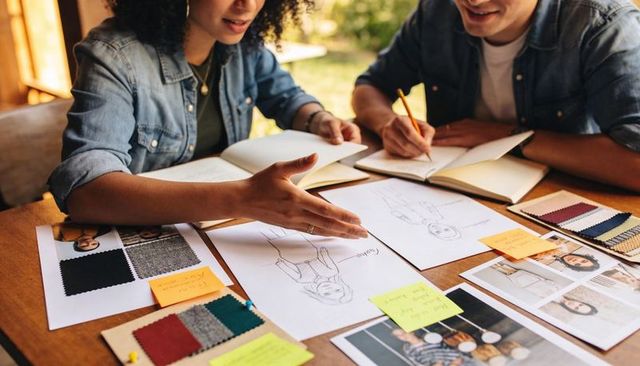 Designers collaborating over fashion sketches and fabric swatches at sunlit workshop table
