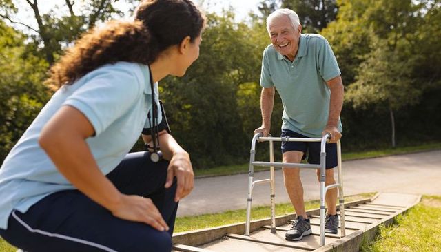 Senior man using walker on balance beam with caregiver assisting outdoor rehabilitation