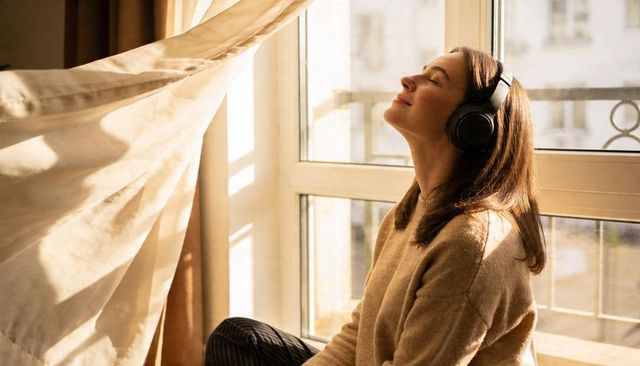 Listening to music on sunlit window sill, woman wearing headphones embracing cozy calm