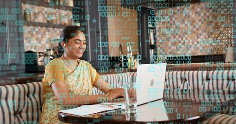 Indian woman typing on laptop in cafe wearing yellow sari, remote work with code overlay