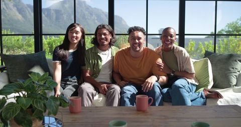 Asian and african american friends relaxing on sofa with mugs in sunlit mountain-view room