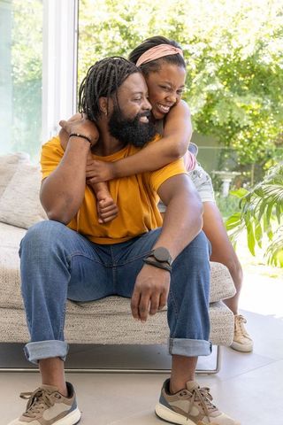 Black father and daughter laughing and hugging on sofa in bright modern home