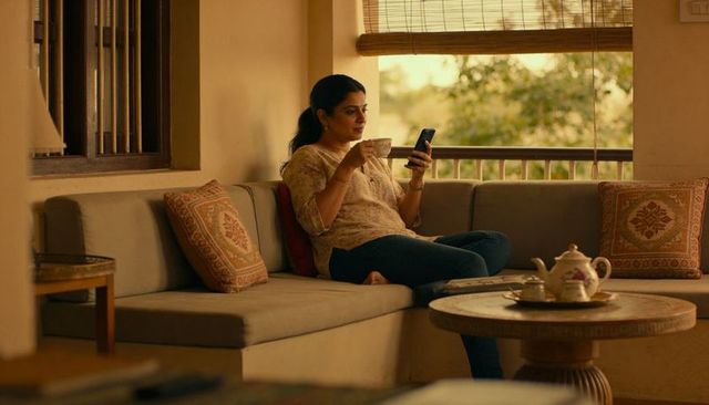 Indian woman relaxing with tea and smartphone on cozy balcony sofa, warm minimalist decor