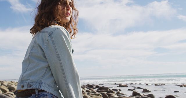 Young woman in denim jacket daydreaming while sitting on rocky beach overlooking ocean under blue sky with clouds. Ideal for use in campaigns focusing on youth, serenity, nature, travel, fashion, casual wear, lifestyle, relaxation, and adventure.