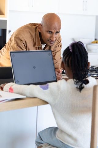 African american father helping child with laptop at kitchen island, home learning support
