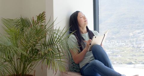Sunlit mindful journaling at window with indoor palm, woman reflecting and writing