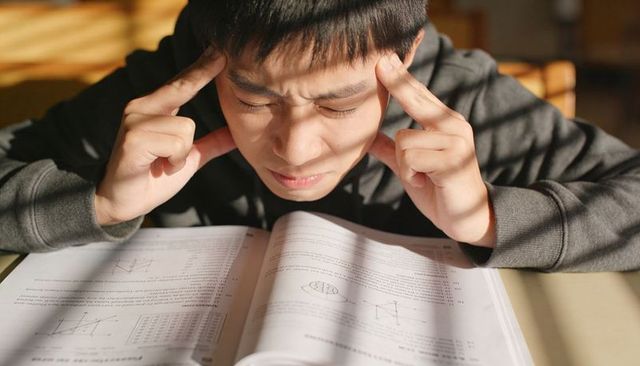 Teen boy pressing temples while studying math textbook under sunlight and shadows