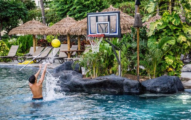 Young man shooting a basketball in a resort pool, splashing water. Tropical setting with lush plants and beach umbrellas. Perfect for illustrating sports, leisure activities, vacation time, and relaxation at exotic locations.