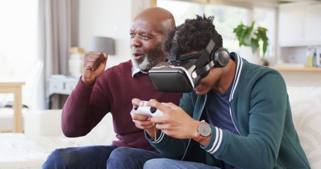 Elderly man cheering while young man wearing VR headset and using game controller. Perfect for illustrating family bonding, generational play, gaming experiences, or the integration of modern technology in everyday life. Can be used in advertisements about virtual reality, family time, interactive technology, and online gaming.