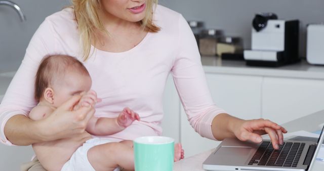 Multitasking Mother Holding Baby and Working on Laptop in Modern Kitchen - Download Free Stock Images Pikwizard.com