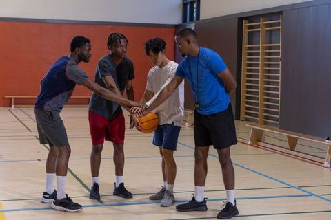 Diverse male basketball teammates huddling midcourt stacking hands on ball before game