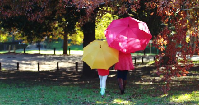 Mother and daughter walking together in an autumn park, holding red and yellow umbrellas. The park is filled with vibrant fall foliage. This image is ideal for promoting family time, seasonal activities, and outdoor bonding moments during the autumn season.