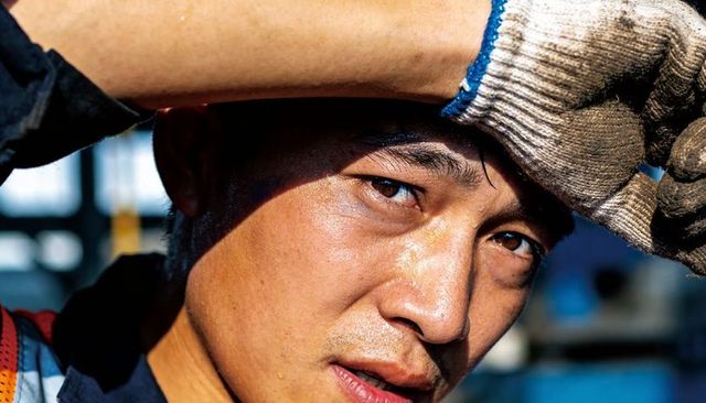 Perspiring asian construction worker shielding eyes with gloved hand on sunlit scaffolding