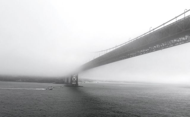 Boat sailing under a large bridge partially obscured by thick fog in a black and white image. The scene captures the misty weather and peaceful waters, creating a serene urban landscape. Ideal for projects on urban transportation, foggy weather conditions, or calmness. Suitable for use in websites, blogs, travel articles, or atmospheric design projects.